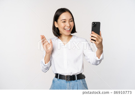 Image of stylish modern asian woman waving hand at smartphone front camera, video chat, chatting with person on mobile phone application, white background Image of stylish modern asian woman waving hand at smartphone front camera, video chat, chatting with person on mobile phone application, white background 86910773