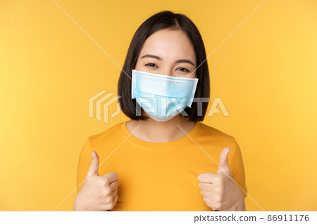 Cheerful korean woman in medical face mask, support people during pandemic, wear personal protective equipment from covid-19, showing thumbs up in approval, yellow background 86911176