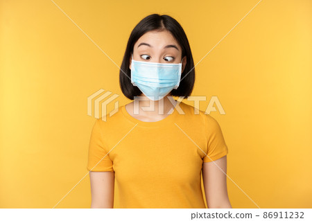 Health and people concept. Funny asian girl squinting, looking at her face medical mask, standing over yellow background Health and people concept. Funny asian girl squinting, looking at her face medical mask, standing over yellow background 86911232