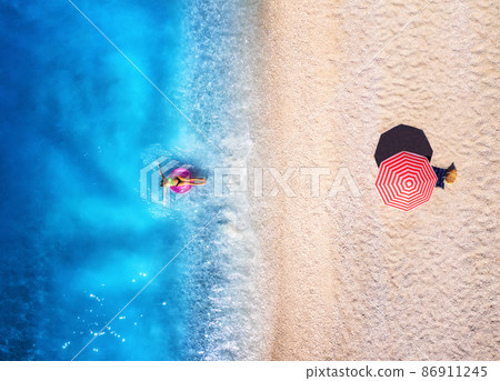 Aerial view of a woman swimming with pink swim ring in blue sea, sandy beach and red umbrella 86911245