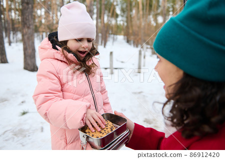 Cute little girl in pink pastel warm winter clothes takes a cookie out of a lunch box and enjoys a break for lunck with her loving mother while resting together in a snowy pine woodland Cute little girl in pink pastel warm winter clothes takes a cookie out of a lunch box and enjoys a break for lunck with her loving mother while resting together in a snowy pine woodland 86912420