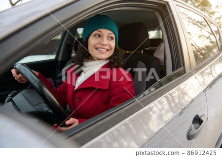 Beautiful woman in bright red coat, white woolen scarf and knit green hat smiles while driving a car. Female driver confidently parking her vehicle on a snow covered woodland Beautiful woman in bright red coat, white woolen scarf and knit green hat smiles while driving a car. Female driver confidently parking her vehicle on a snow covered woodland 86912425