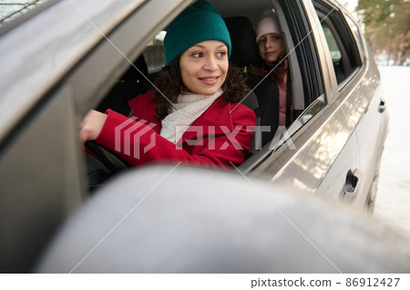 View through blurred driver's side mirror of adorable happy driver mom traveling with kids by car on snow covered road on a beautiful winter snowy day. Winter holidays, adventure, discover nature View through blurred driver's side mirror of adorable happy driver mom traveling with kids by car on snow covered road on a beautiful winter snowy day. Winter holidays, adventure, discover nature 86912427