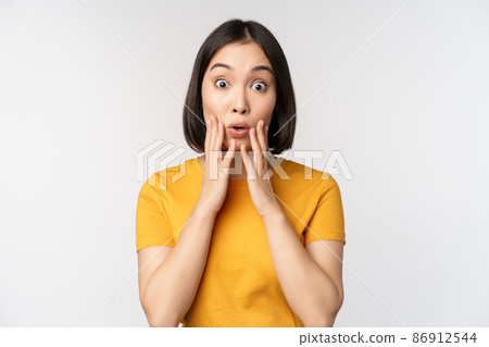 Close up portrait of asian woman looking surprised, wow face, staring impressed at camera, standing over white background in yellow t-shirt 86912544