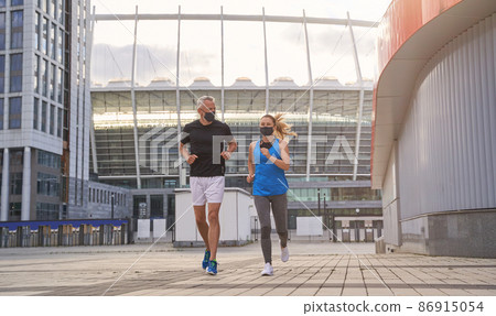 Full length shot of middle aged healthy couple in protective masks jogging together in the morning in the city Full length shot of middle aged healthy couple in protective masks jogging together in the morning in the city 86915054