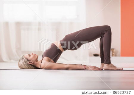 Attractive blonde woman in sports beige leggings and tank top, practicing yoga, doing Glute bridge exercise, dvi pada pithasana pose, home workout 86923727