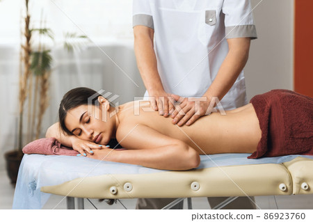Young beautiful brunette woman is resting in the spa salon at the reception of the masseur, the specialist performs a massage of the back muscles 86923760