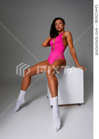 Beautiful sexy woman posing in pink fashionable swimsuit with smile on gray background in a studio on a cube, looking at camera Beautiful sexy woman posing in pink fashionable swimsuit with smile on gray background in a studio on a cube, looking at camera 86925445