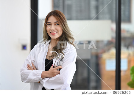 Confident asian female doctor in white uniform standing in medical office and smiling at camera. Confident asian female doctor in white uniform standing in medical office and smiling at camera. 86926265