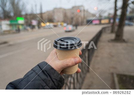 A hand holds a paper cup of coffee against blurry city traffic. A grown man with a hot beverage on the street. Selective focus A hand holds a paper cup of coffee against blurry city traffic. A grown man with a hot beverage on the street. Selective focus 86926282