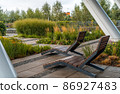 Two empty brown wooden deck chairs or chaise longues on tile among decorative grass and flowers in recreation area. Garden landscape with chairs in city park. Concept of recreation, tanning in yard 86927483