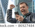 Excited guy looks at phone and celebrates big win financial good news success achievement cell phone in front of modern buildings. Happy man checking news on smart phone walking in street 86927484
