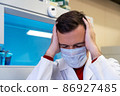 Millennial guy suffering migraine headache problem holding head with hands. Middle age professional scientist man wearing white coat with hand on head for pain in head because stress in laboratory 86927485