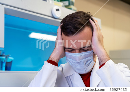 Millennial guy suffering migraine headache problem holding head with hands. Middle age professional scientist man wearing white coat with hand on head for pain in head because stress in laboratory Millennial guy suffering migraine headache problem holding head with hands. Middle age professional scientist man wearing white coat with hand on head for pain in head because stress in laboratory 86927485