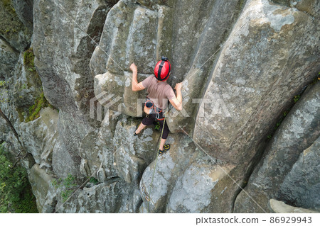 Young man climbing steep wall of rocky mountain. Male climber overcomes challenging route. Engaging in extreme sport concept. 86929943