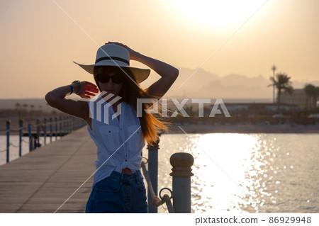 Young happy woman tourist relaxing outdoors on warm sunny evening on sea shore. Summer vacations and travelling concept. Young happy woman tourist relaxing outdoors on warm sunny evening on sea shore. Summer vacations and travelling concept. 86929948