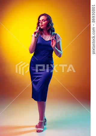 Emotional young girl with long curly hair in retro style attire isolated on orange background in neon light, filter. Concept of emotions, beauty, fashion 86930801