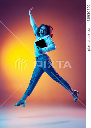 Full-length portrait of happy young beautiful girl with long curly hair jumping isolated on orange background in neon light, filter. Concept of emotions, beauty, fashion 86930802