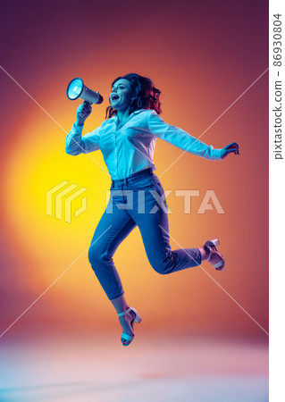 Full-length portrait of happy young beautiful girl with long curly hair jumping isolated on orange background in neon light, filter. Concept of emotions, beauty, fashion Full-length portrait of happy young beautiful girl with long curly hair jumping isolated on orange background in neon light, filter. Concept of emotions, beauty, fashion 86930804