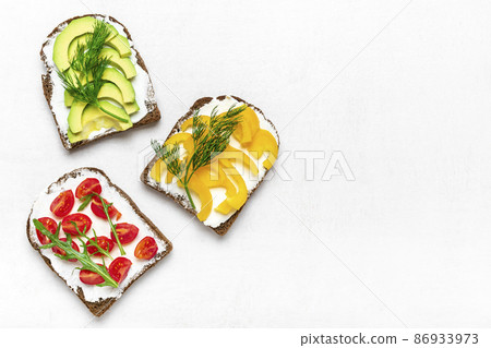 Variety of sandwiches for breakfast - slice of whole grain dark bread, yellow bell pepper, cream cheese, avocado, cherry tomatoes, garnished with dill, green arugula on white table Top view Flat lay Variety of sandwiches for breakfast - slice of whole grain dark bread, yellow bell pepper, cream cheese, avocado, cherry tomatoes, garnished with dill, green arugula on white table Top view Flat lay 86933973