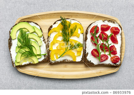 Variety of sandwiches for breakfast - slice of whole grain dark bread, yellow bell pepper, cream cheese, avocado, cherry tomatoes, garnished with dill, green arugula on table Top view Flat lay 86933976