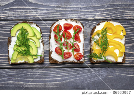 Variety of sandwiches for breakfast - slice of whole grain dark bread, yellow bell pepper, cream cheese, avocado, cherry tomatoes, garnished with dill, green arugula on wooden table Top view Flat lay Variety of sandwiches for breakfast - slice of whole grain dark bread, yellow bell pepper, cream cheese, avocado, cherry tomatoes, garnished with dill, green arugula on wooden table Top view Flat lay 86933997