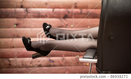 Women's slender legs in shoes lie on the armrest of an office chair against the background of a wooden wall with a garland Women's slender legs in shoes lie on the armrest of an office chair against the background of a wooden wall with a garland 86938271