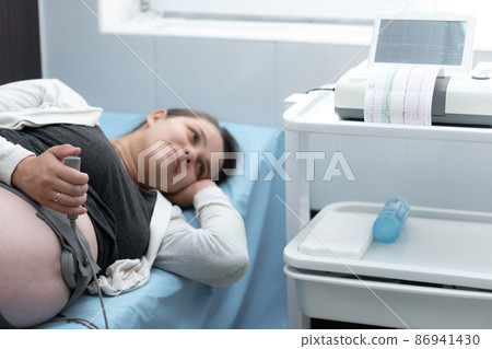 Pregnant Woman On Hospital Couch During Medical Control Cardiotocography. Gynecologist Doctor Hands Turn On Adjust CTG Device Clinic Consultant. Examine Expectant Belly Baby Mother Healthcare Check Up 86941430
