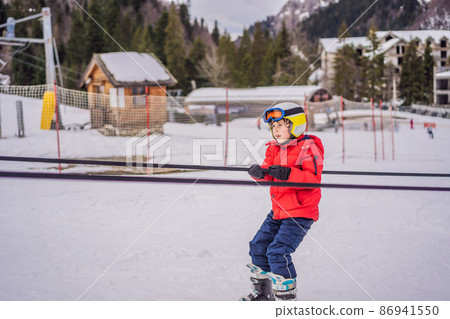 Boy uses a training lift. Child skiing in mountains. Active toddler kid with safety helmet, goggles and poles. Ski race for young children. Winter sport for family. Kids ski lesson in alpine school Boy uses a training lift. Child skiing in mountains. Active toddler kid with safety helmet, goggles and poles. Ski race for young children. Winter sport for family. Kids ski lesson in alpine school 86941550