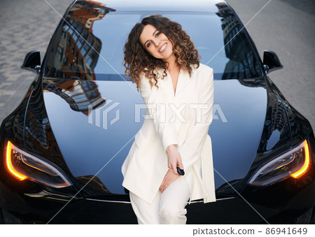 Half length portrait of cheerful female entrepreneur dressed in white suit smiling at camera while putting lock button on car key, successful Caucasian businesswoman posing near black electric car. Half length portrait of cheerful female entrepreneur dressed in white suit smiling at camera while putting lock button on car key, successful Caucasian businesswoman posing near black electric car. 86941649