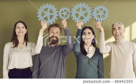 Team of four creative business people connecting gear wheels. Studio group portrait of 4 happy smiling adult men and women joining cogwheels to show concept of effective teamwork and collaboration 86942393