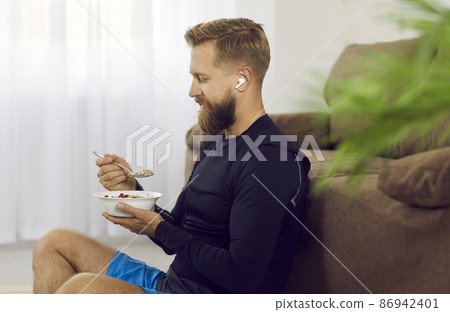 Athletic man after active morning sports training eats healthy food to restore energy. Side view of bearded man with wireless headphones in his ears sitting on floor near sofa with bowl of oatmeal. 86942401