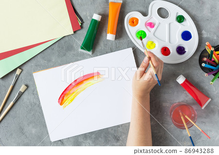 Woman hand painting rainbow in sketchbook with pain brush with art supplies around top view on grey concrete table. 86943288