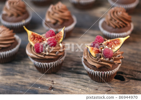 Delicious chocolate muffins with butter cream, raspberries and fig pieces. Coffee cupcakes on wooden table, close-up 86944970