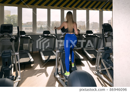 Back view on young beautiful girl with long hair trains in the gym. Elliptical trainer for cardio. 86946096