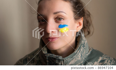 Indoor portrait of young girl with blue and yellow ukrainian flag on her cheek wearing military uniform, mandatory conscription in Ukraine, equality concepts Indoor portrait of young girl with blue and yellow ukrainian flag on her cheek wearing military uniform, mandatory conscription in Ukraine, equality concepts 86947104