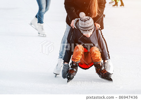 Soft,Selective focus.Mom with baby boy 6 years old, learn train, ride winter city rink, ice skating. The child gets up, fell on skates, kneels, play fun rest on weekend first steps child skates Soft,Selective focus.Mom with baby boy 6 years old, learn train, ride winter city rink, ice skating. The child gets up, fell on skates, kneels, play fun rest on weekend first steps child skates 86947436