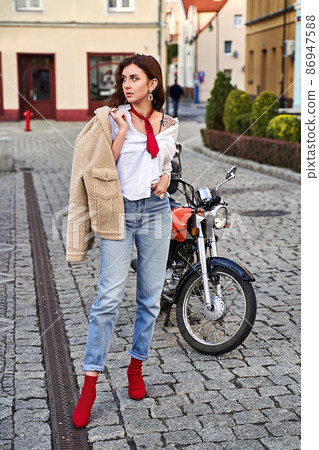 Young beautiful woman in red knitted fabric block heel sock shoes, blue denim jeans pants and teddy jacket coat posing near a motorcycle on a street Young beautiful woman in red knitted fabric block heel sock shoes, blue denim jeans pants and teddy jacket coat posing near a motorcycle on a street 86947588