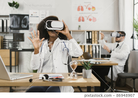 Multiethnic medical workers in VR headset sitting at cabinet Multiethnic medical workers in VR headset sitting at cabinet 86948907