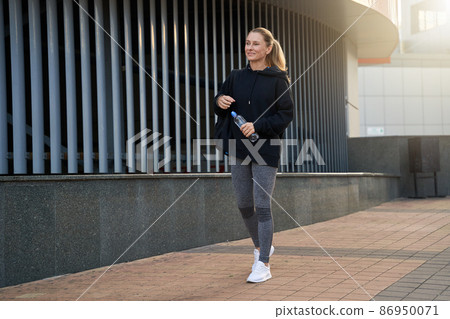 Full length shot of attractive middle aged sportswoman in sportswear smiling while walking outdoors, ready for workout Full length shot of attractive middle aged sportswoman in sportswear smiling while walking outdoors, ready for workout 86950071