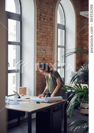 Interior designer or architect in casual wear with messy hairdo looking focused while working on a blueprint for new project, standing in her office on a daytime 86958266