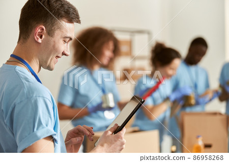 Side view of happy male volunteer smiling, using tablet pc while working on donation project with team. Focus on man 86958268
