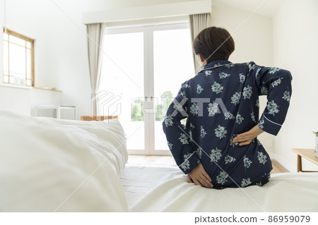 Back view of a middle-aged woman in pajamas sitting on the bed and holding her hips Back view of a middle-aged woman in pajamas sitting on the bed and holding her hips 86959079