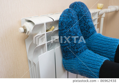 A woman warms her feet by the radiator on a cold winter day in blue woolen socks. Central heating system. Expensive heating costs during the cold season A woman warms her feet by the radiator on a cold winter day in blue woolen socks. Central heating system. Expensive heating costs during the cold season 86962438