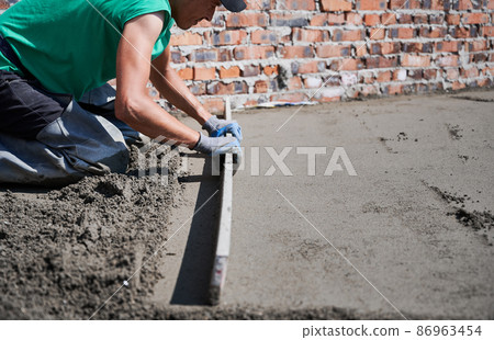 Close up of man builder placing screed rail on the floor covered with sand-cement mix at construction site on the roof. Male worker leveling surface with straight edge while screeding floor. Close up of man builder placing screed rail on the floor covered with sand-cement mix at construction site on the roof. Male worker leveling surface with straight edge while screeding floor. 86963454