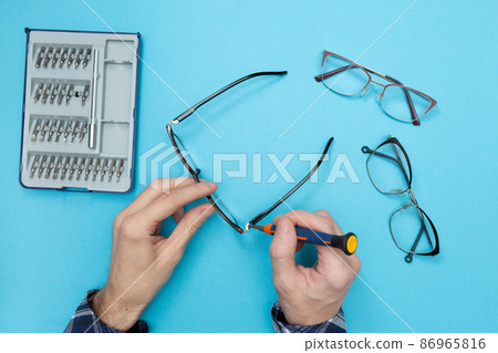Master uses a screwdriver to tighten the screw in the frame of the glasses. Eyeglass frame repair. Optics salon and repair shop. Flat lay. Blue background. Master uses a screwdriver to tighten the screw in the frame of the glasses. Eyeglass frame repair. Optics salon and repair shop. Flat lay. Blue background. 86965816