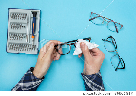 Man wiping glasses for vision with a napkin. Cleaning glasses. Glasses repair workshop. Blue background. Flat lay. Man wiping glasses for vision with a napkin. Cleaning glasses. Glasses repair workshop. Blue background. Flat lay. 86965817