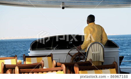 Experienced Yacht Captain at steering wheel controls sailing motor boat. Senior man in yellow uniform turns helm on modern ship dashboard in blue sea. Strong wind makes waves, vessel rocks. Sea voyage 86967188