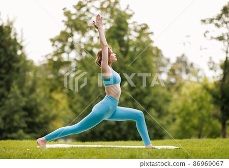 Slender focused girl performing yoga outside in early morning in park, standing in warrior pose Slender focused girl performing yoga outside in early morning in park, standing in warrior pose 86969067