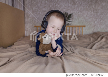 Boy in headphones listens to a meditation for children before going to bed. Concept of a healthy nervous system of children. Relaxation before sleep. Blond guy is relaxing at home. Hearing therapy Boy in headphones listens to a meditation for children before going to bed. Concept of a healthy nervous system of children. Relaxation before sleep. Blond guy is relaxing at home. Hearing therapy 86969370